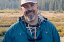 A man with a gray beard and cap smiles outdoors, standing in front of a field and forest with mountains in the background.