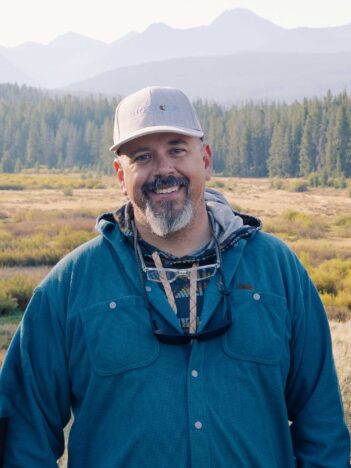 A man with a gray beard and cap smiles outdoors, standing in front of a field and forest with mountains in the background.
