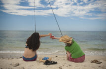 Two people sitting on a sandy beach, fishing and clinking cans together, facing the ocean under a partly cloudy sky.