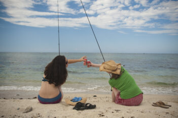 Two people sitting on a sandy beach, fishing and clinking cans together, facing the ocean under a partly cloudy sky.