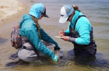 Two people in outdoor gear kneel in shallow water; one holds a fish while the other writes on a notepad.